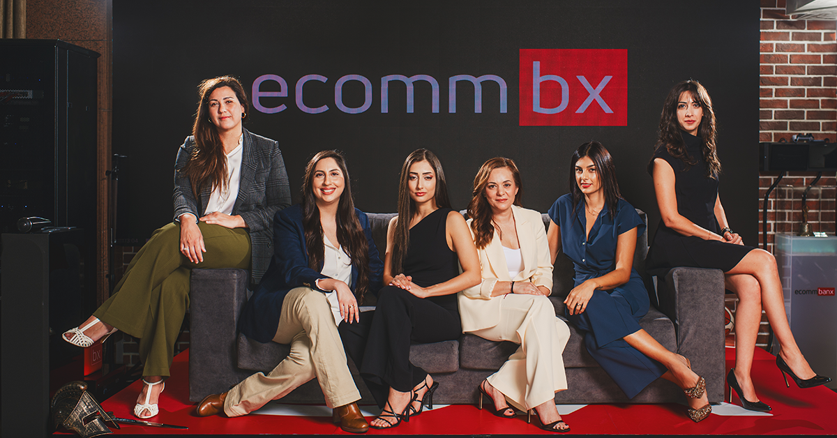Seven women in business attire pose on a gray sofa in front of an ecommbx backdrop, creating a team portrait.